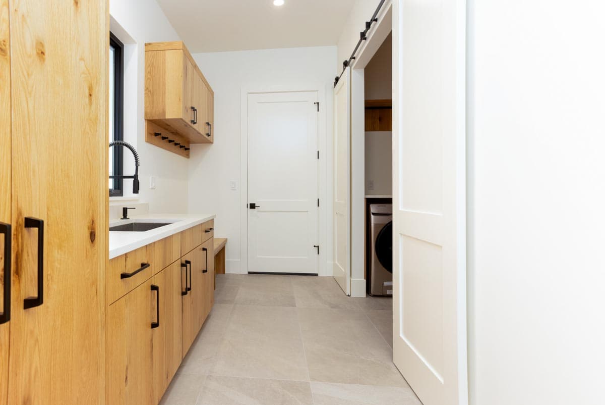 A modern laundry room with light wood cabinetry, a sink, and a white countertop. A sliding door reveals a washing machine.