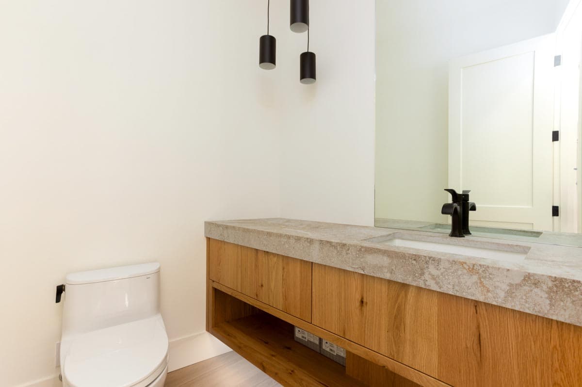 Modern bathroom with wooden vanity and concrete countertop. Black faucet and pendant lights are featured, along with a white toilet and mirror.