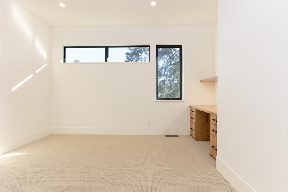 An empty, modern room with light-colored carpet and walls. Windows offer a view of trees. A wooden desk with drawers is to the right.