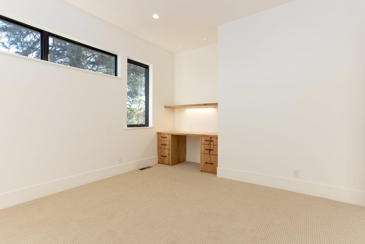An empty, minimalist home office featuring a wooden desk with drawers and a small shelf with lighting against a white wall.