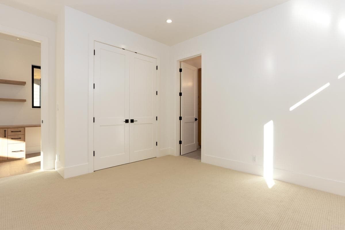 A bright, empty room with white walls, carpeted floor, and two closed closet doors. An open doorway leads to another room.