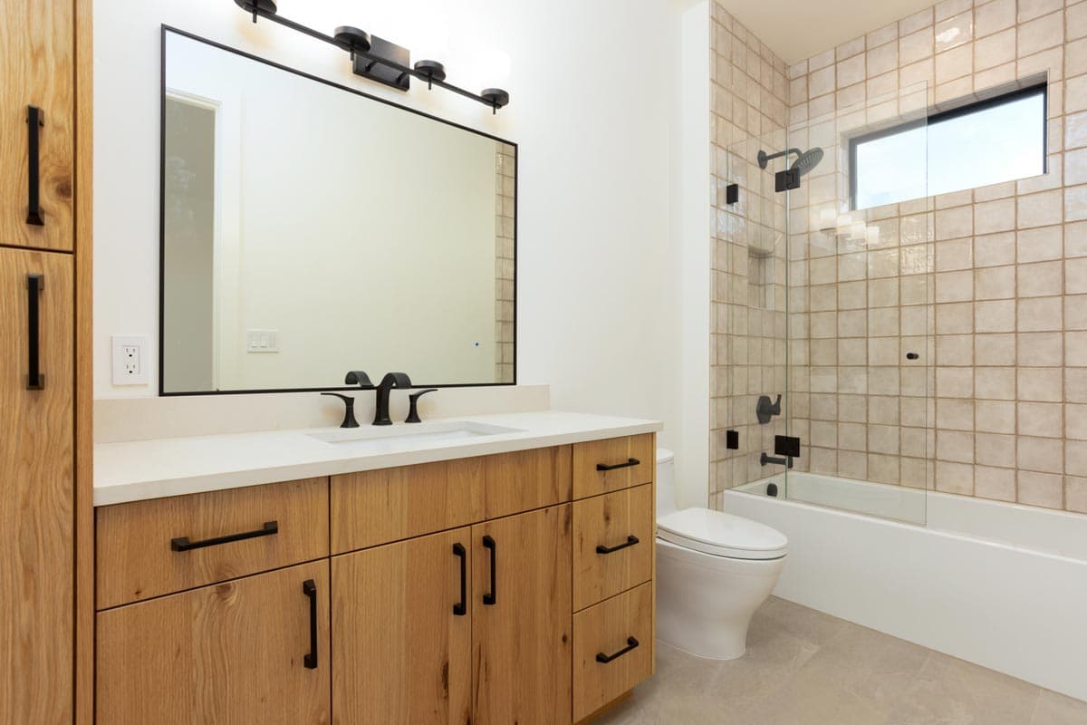 A modern bathroom features a wooden vanity with black hardware, a large mirror, and a tiled shower with a glass door and a small window.