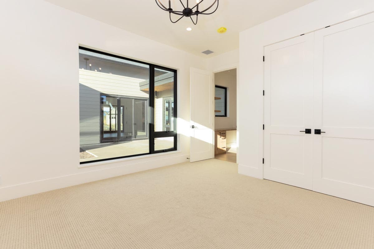 An empty room with beige carpet, a large window, and white walls. A door is ajar, and a black light fixture hangs from the ceiling.