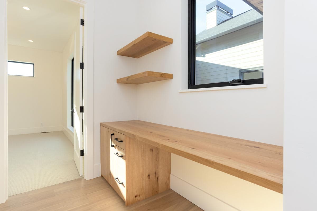 Modern office space with a built-in wooden desk, floating shelves, and a large window overlooking a house exterior and a door in the background.