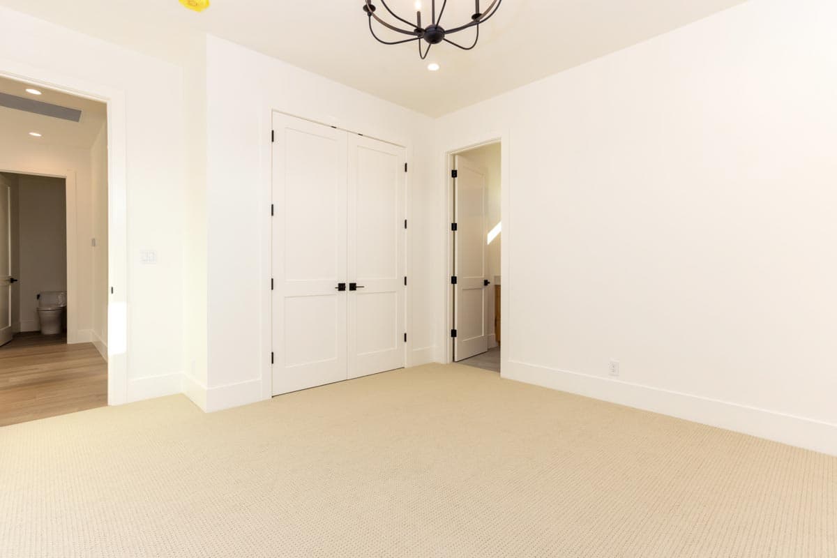 A modern, empty room with beige carpet, white walls, and two closed, white doors with black handles. An open doorway leads to a hallway.