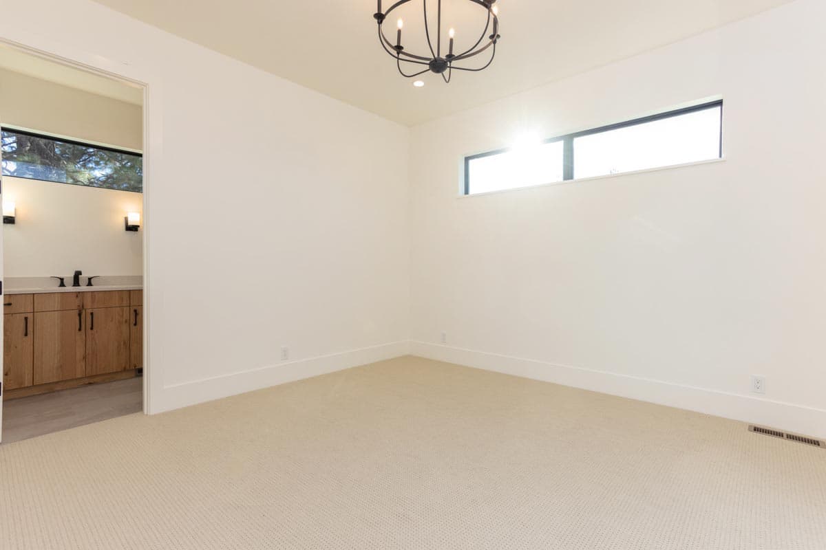 An empty, modern room with off-white walls, carpeted floor, and a chandelier. A doorway leads to a bathroom with wooden cabinets.