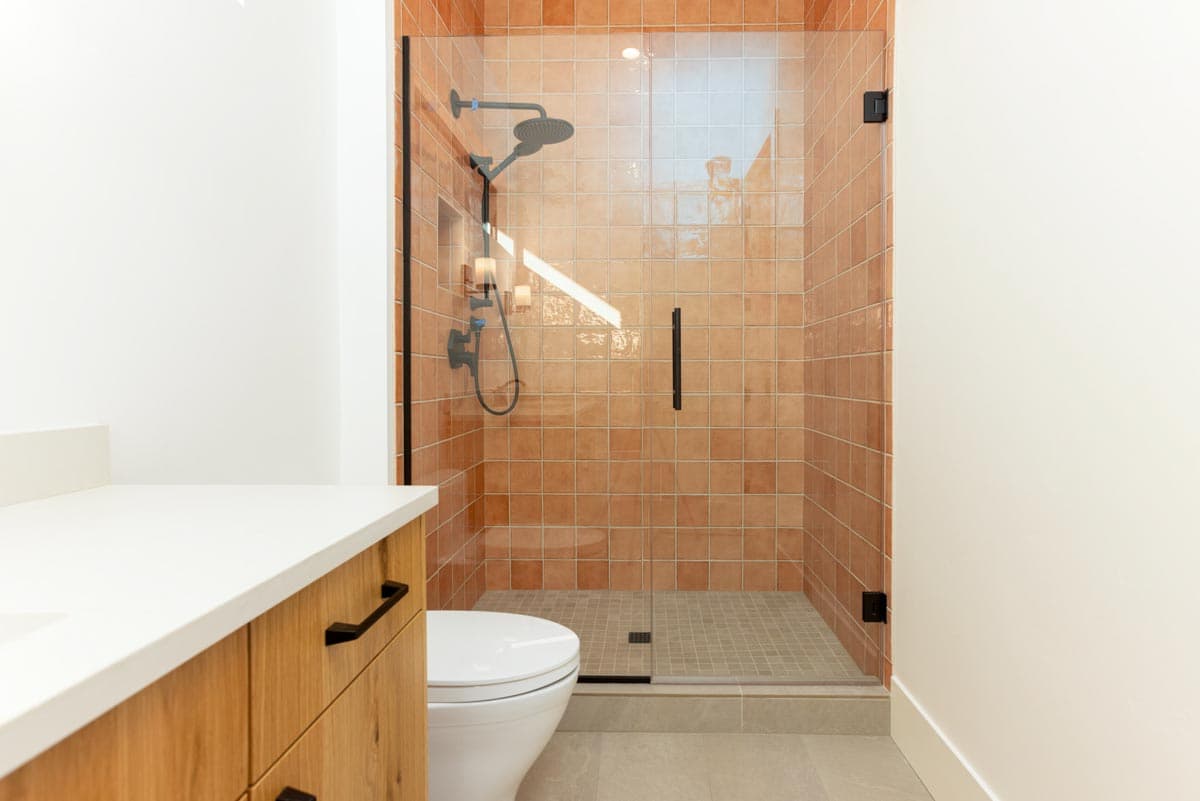 A modern bathroom showcases a walk-in shower with terracotta-colored tiles, glass doors, and black fixtures. A wooden vanity and toilet are in the foreground.