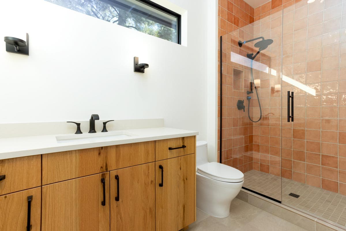Modern bathroom with a wooden vanity, a white toilet, and a glass-enclosed shower with orange-tiled walls and a rainfall showerhead.