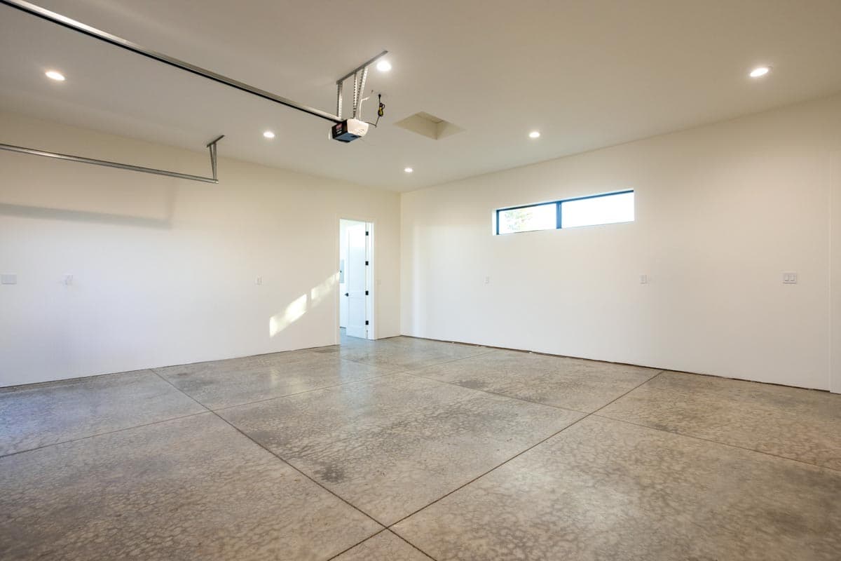 An empty, modern garage with a concrete floor and white walls. A small window lets in light, and a door is slightly ajar in the background.