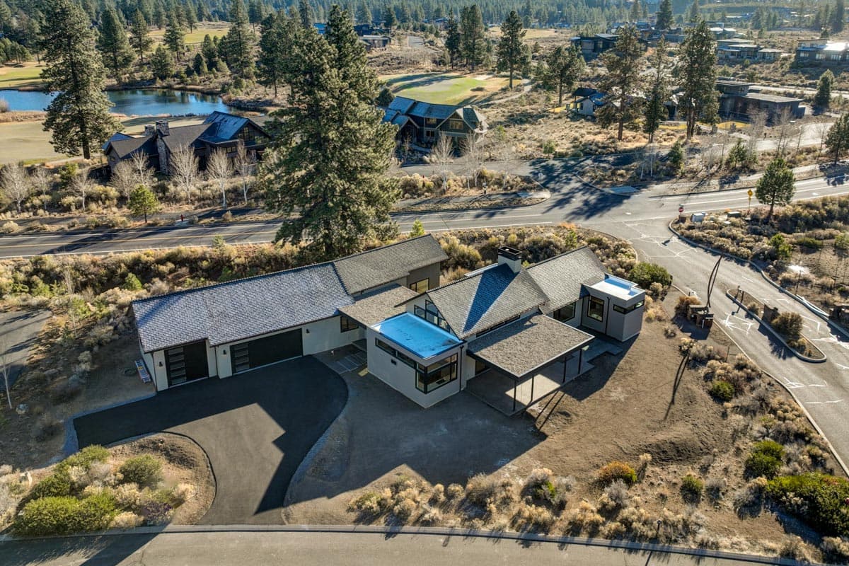 Aerial view of a modern home with a dark gray roof. In the background are other homes, a golf course, and a road winding through a landscape.