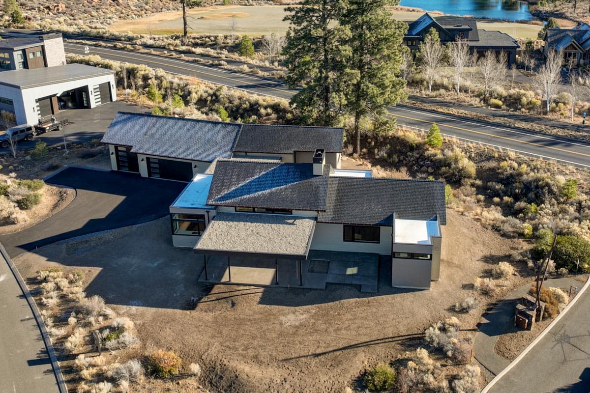 Aerial view of a modern house with a complex roof design, surrounded by dry, shrub-covered landscape. A nearby road and other buildings are visible.
