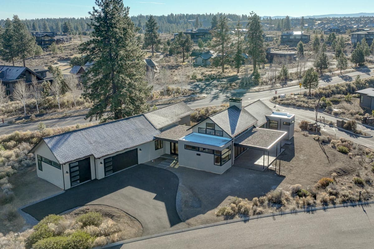 An aerial view of a modern, single-story house with a long driveway and a flat, dark roof. The home is surrounded by roads, trees, and other houses.
