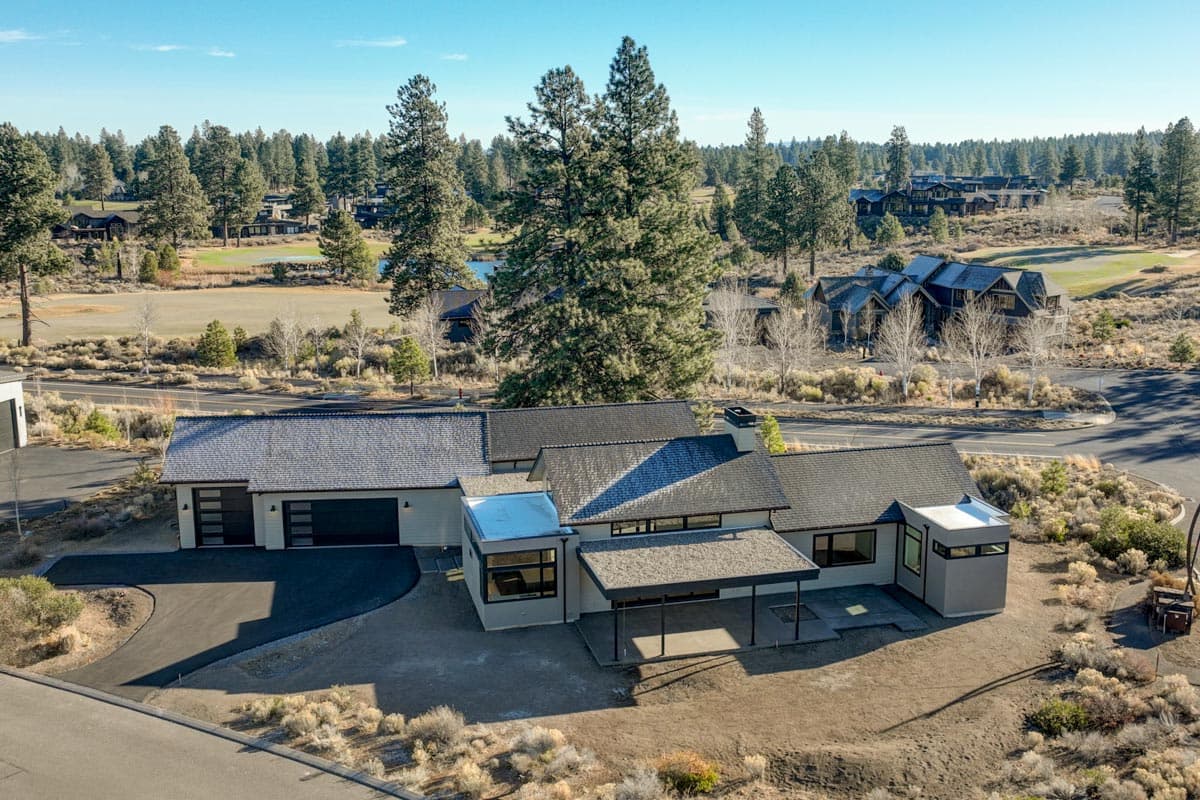 Aerial view of a modern, single-story house with a flat roof, surrounded by a golf course and pine trees under a clear blue sky.