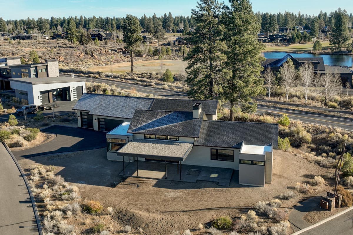An aerial view of a modern house with a dark roof surrounded by dry landscape and trees. A lake and other homes are visible in the distance.
