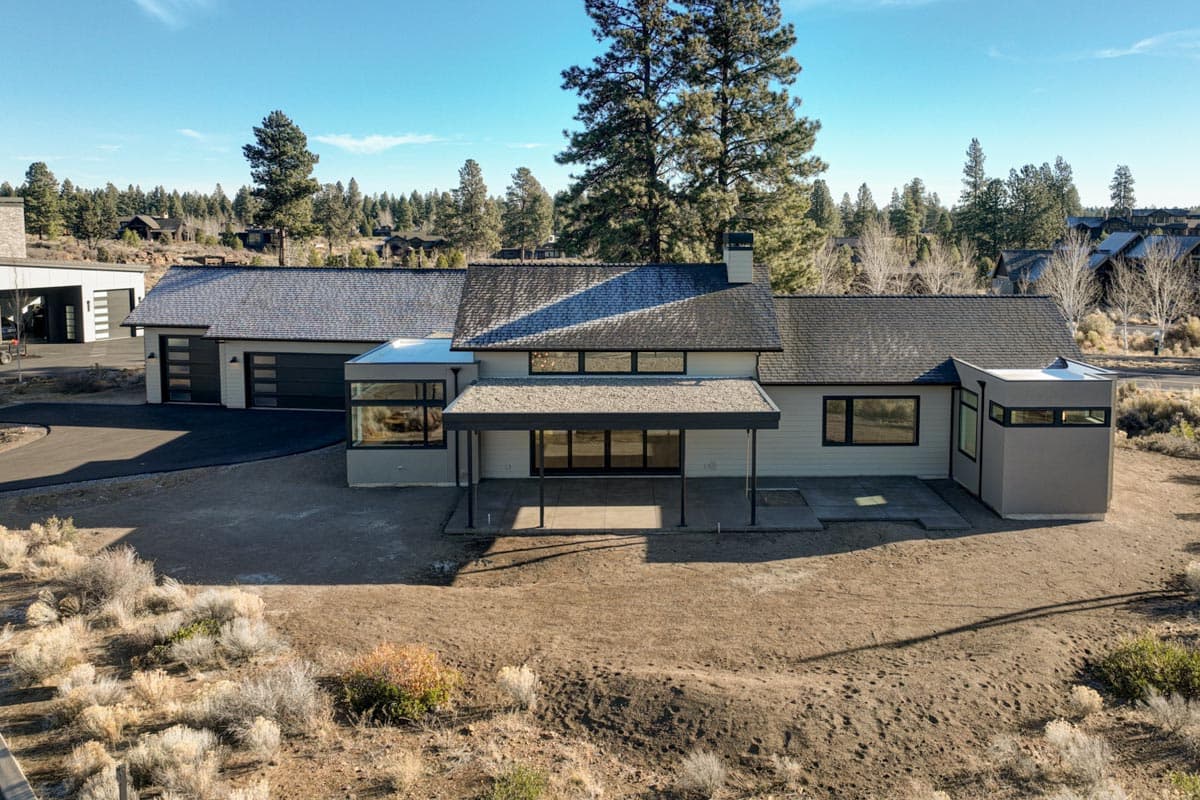 An aerial view showcases a modern, single-story house with a long, gray roof and expansive windows, set against a backdrop of trees.