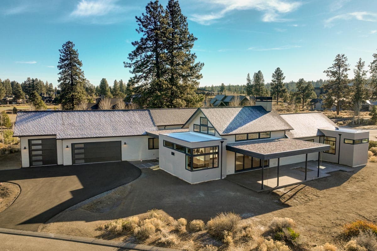 Modern, multi-level house with a gray exterior, black garage doors, and a covered patio. Surrounded by trees and a clear blue sky.