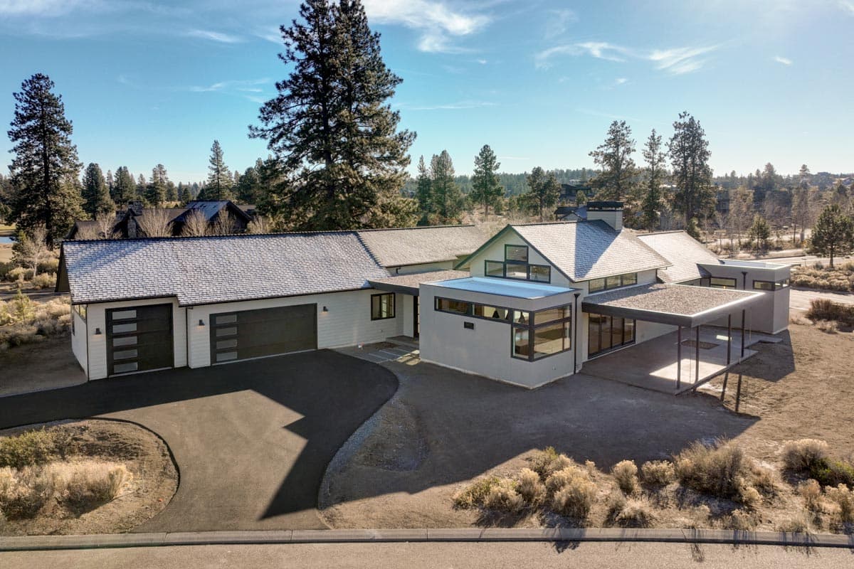 An aerial view of a modern, single-story house with a light gray exterior, a black garage door, and a long driveway leading to it.