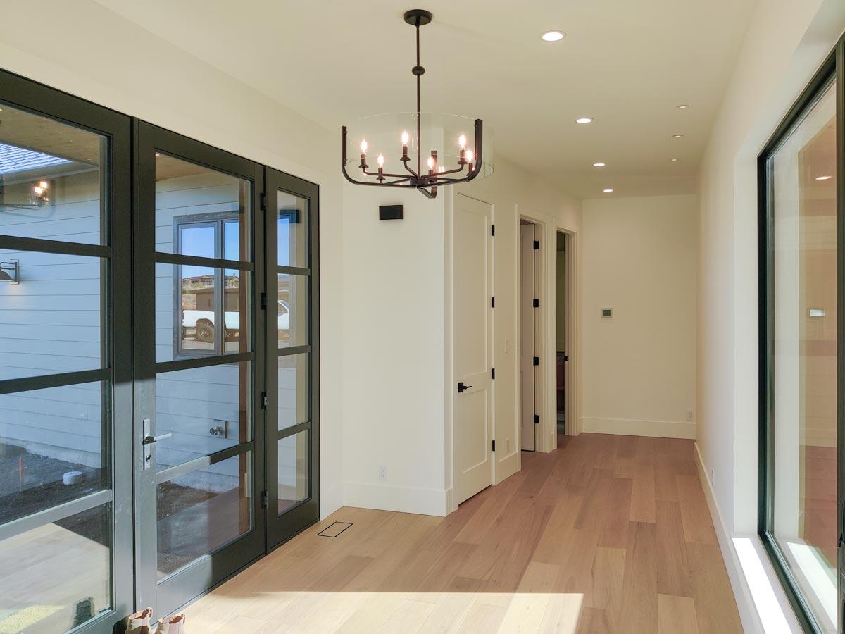 A hallway with light wood floors, white walls, and a decorative chandelier. Dark-framed glass doors are visible on the left, leading to an outside view.