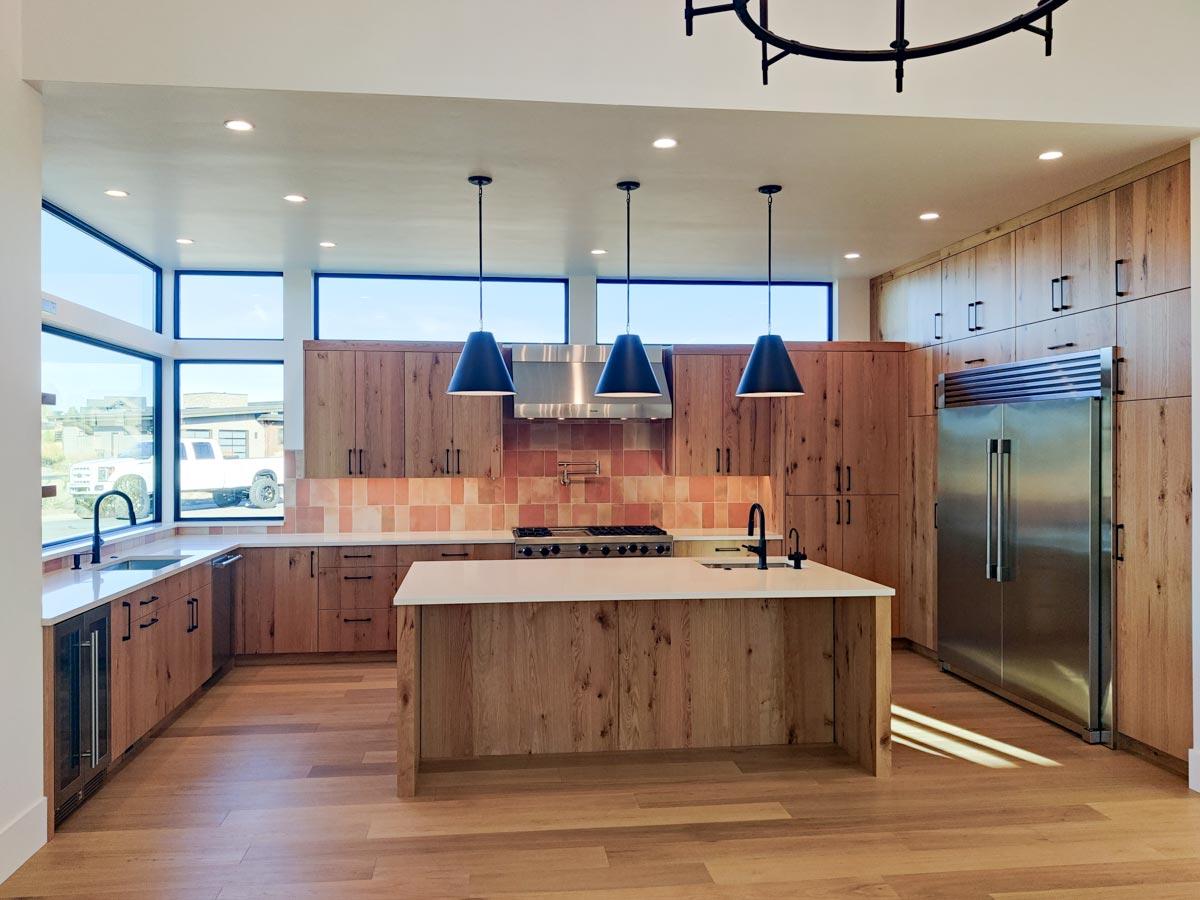Modern kitchen with wood cabinets, island, and large windows. Black pendant lights hang over the stove and a stainless steel refrigerator is present.
