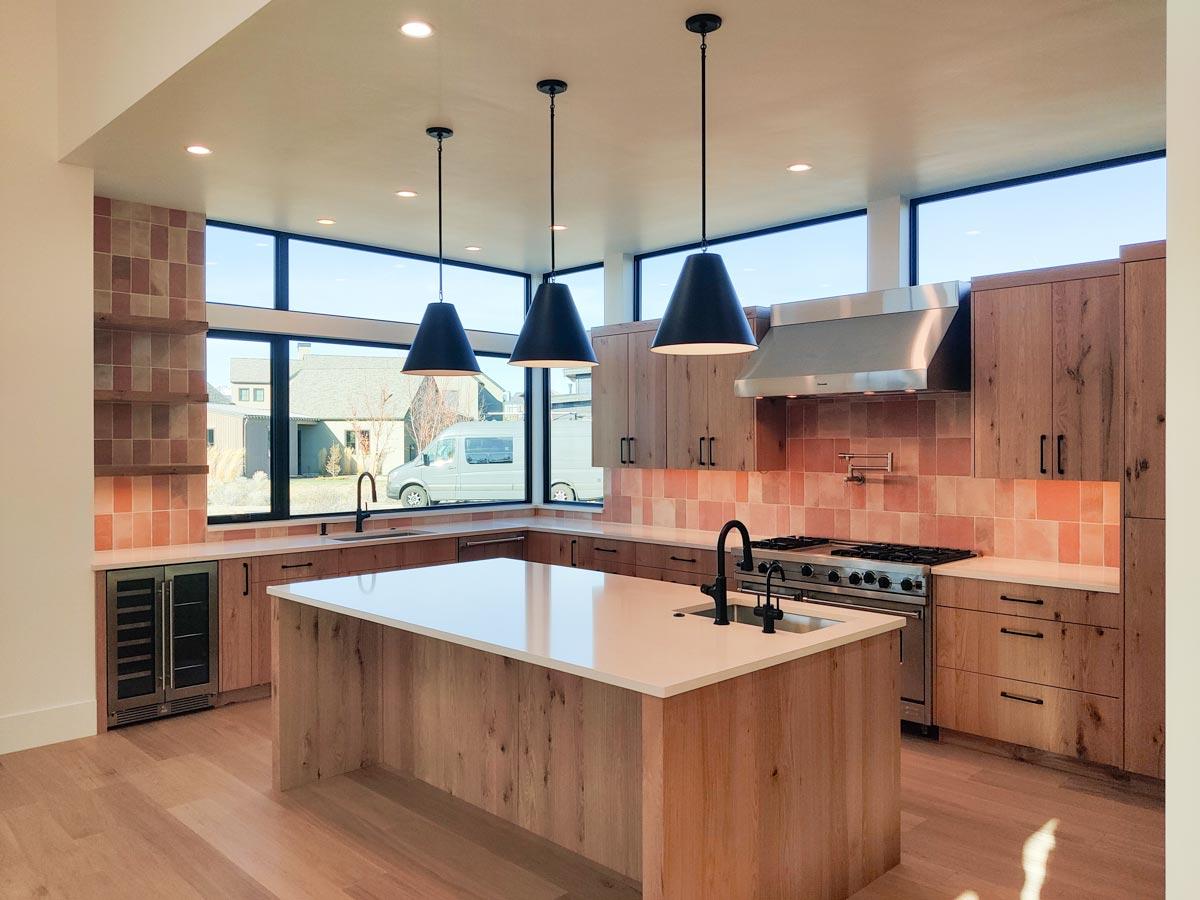 Modern kitchen with wood cabinets and island. Black pendant lights hang above the island. Large windows with a view are in the background.