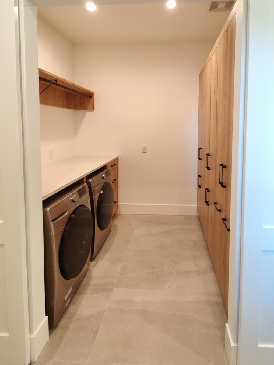A modern laundry room with two silver washing machines, a white countertop, wooden cabinets, and pale gray tiled floor.