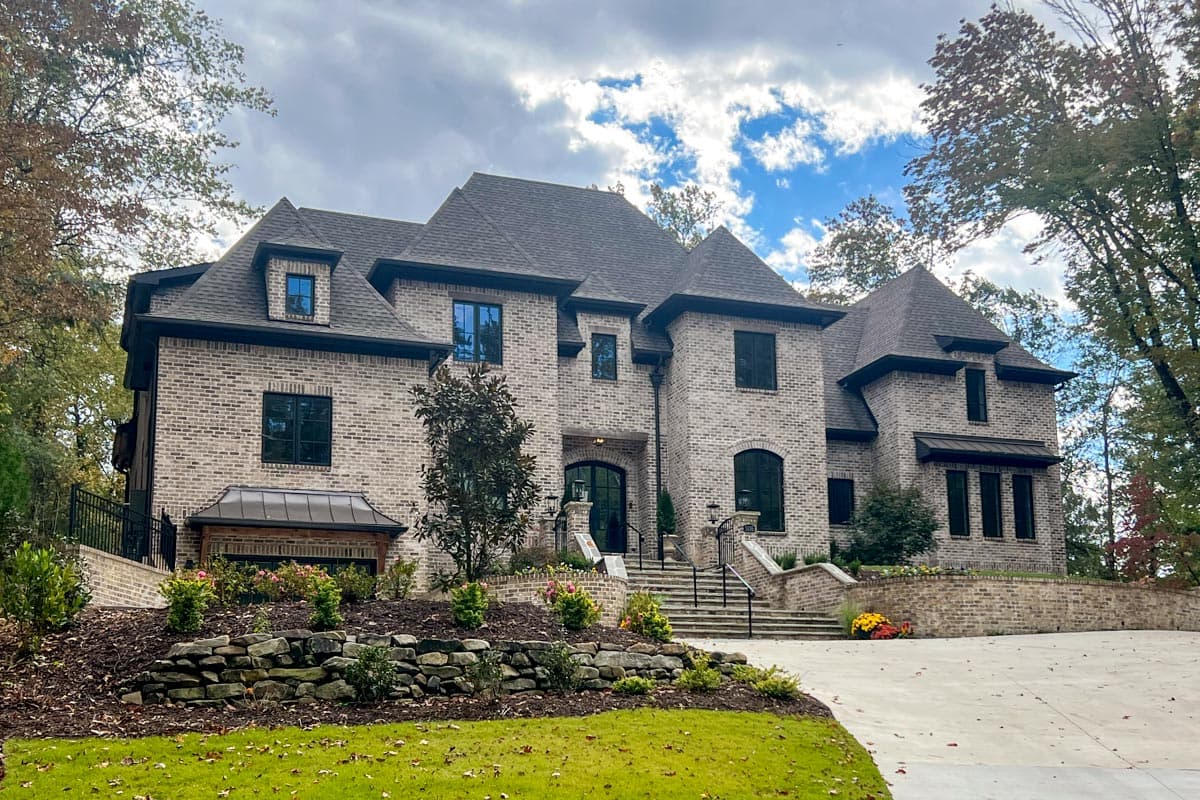 House plan exterior. Two-story brick home with multiple gables, dormers, and a prominent entryway. Includes landscaping and a tiered driveway.