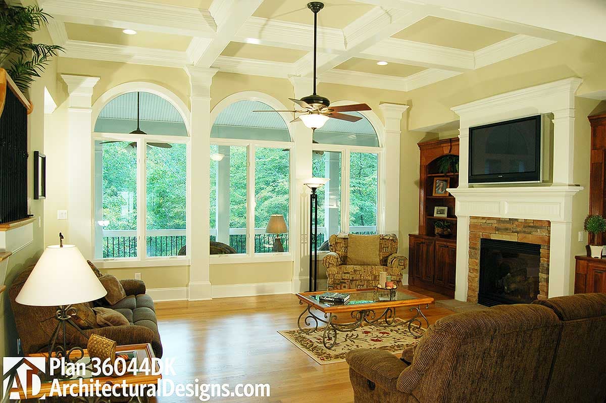 Living room with coffered ceiling, fireplace, built-in cabinetry, and large arched windows overlooking wooded area.