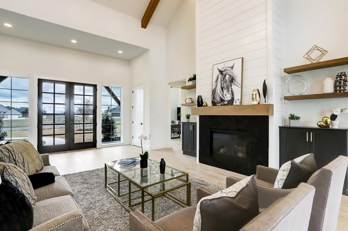 A modern living room with large windows, fireplace, and two armchairs. A horse portrait hangs above the fireplace, with shelves on the wall.