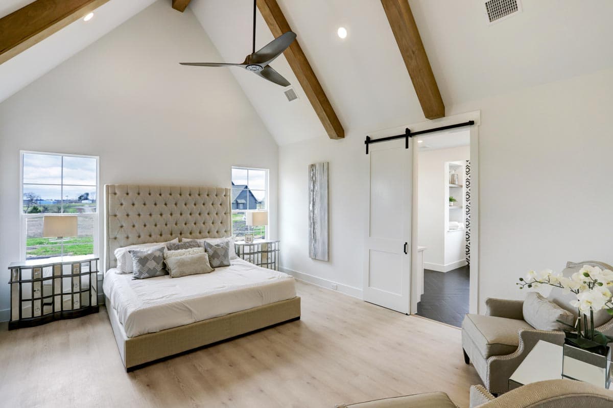 A luxurious bedroom with a high vaulted ceiling, exposed wooden beams, and a large tufted headboard. A sliding door reveals a glimpse of a bathroom.