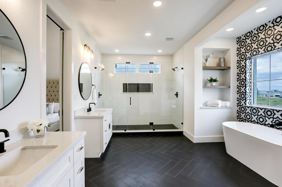 Modern bathroom with white walls, black herringbone floor, glass shower, and freestanding tub beside a decorative tile wall and window.