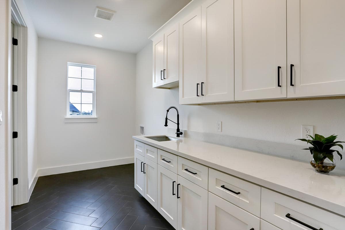 A modern laundry room featuring white cabinetry, a dark herringbone floor, and a black faucet. A small window lets in natural light.