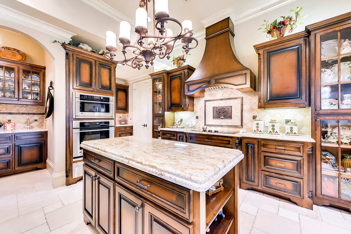 Kitchen interior with central island, dark wood cabinetry, stacked ovens, ornate chandelier, and decorative range hood.