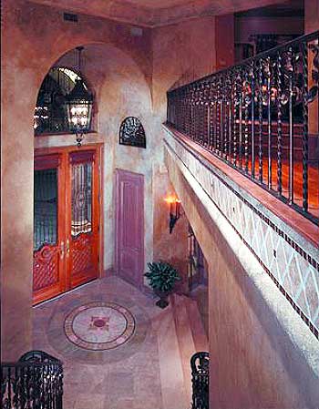 Interior view of a grand foyer with double doors, arched entryway, decorative floor inlay, and wrought iron railing on upper level.