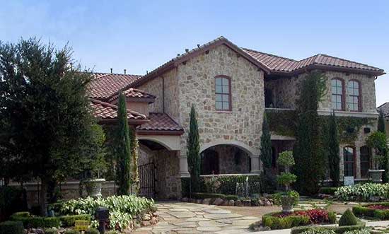 Two-story stone house plan with arched entryway, tile roof, numerous windows, and manicured landscaping.