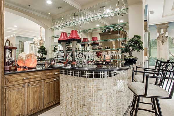 Home bar with mosaic tile base, wooden cabinetry, glass shelving displaying barware, and two counter stools.