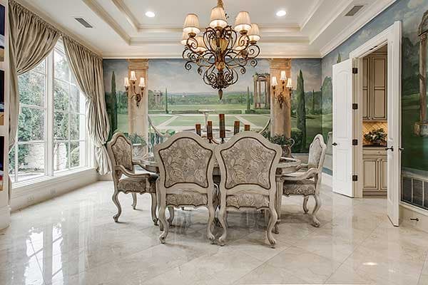 Dining room with a large chandelier, upholstered chairs, a table, and a mural depicting an outdoor landscape. Features large windows.
