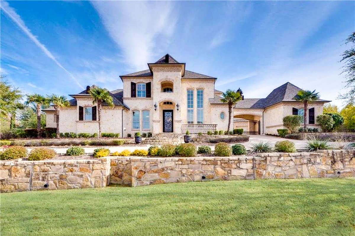 House plan exterior with two stories, arched entryway, gabled rooflines, and an attached garage. Stone wall and landscaping in the foreground.