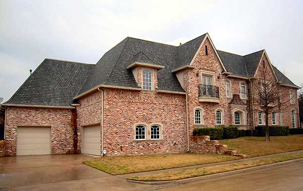 House plan exterior: Traditional brick two-story with side-entry garages, multiple gables, dormers, and a decorative balcony.