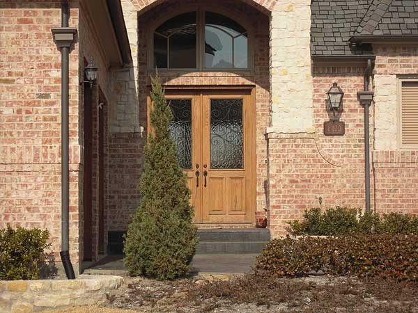 House plan exterior featuring double wooden doors with glass, arched window, brick facade, and stone accents.