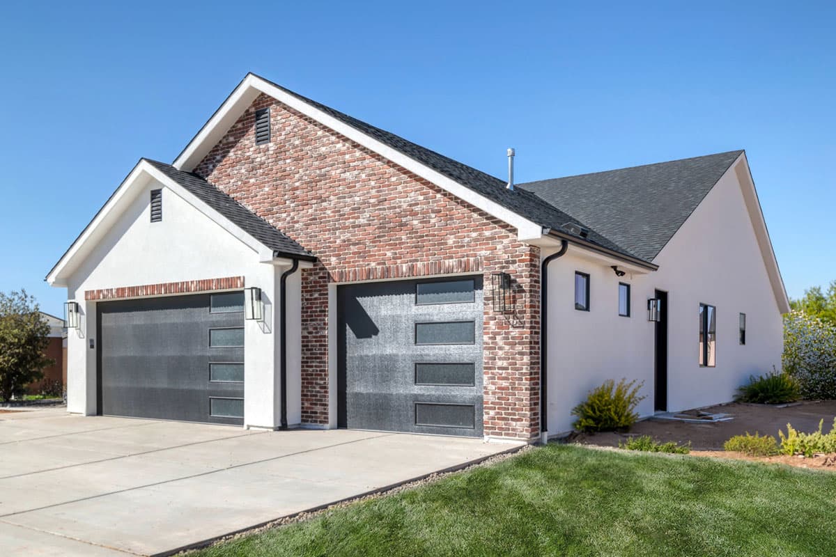 House plan exterior with stucco, brick accents, gable rooflines, and a two-car garage with modern doors.