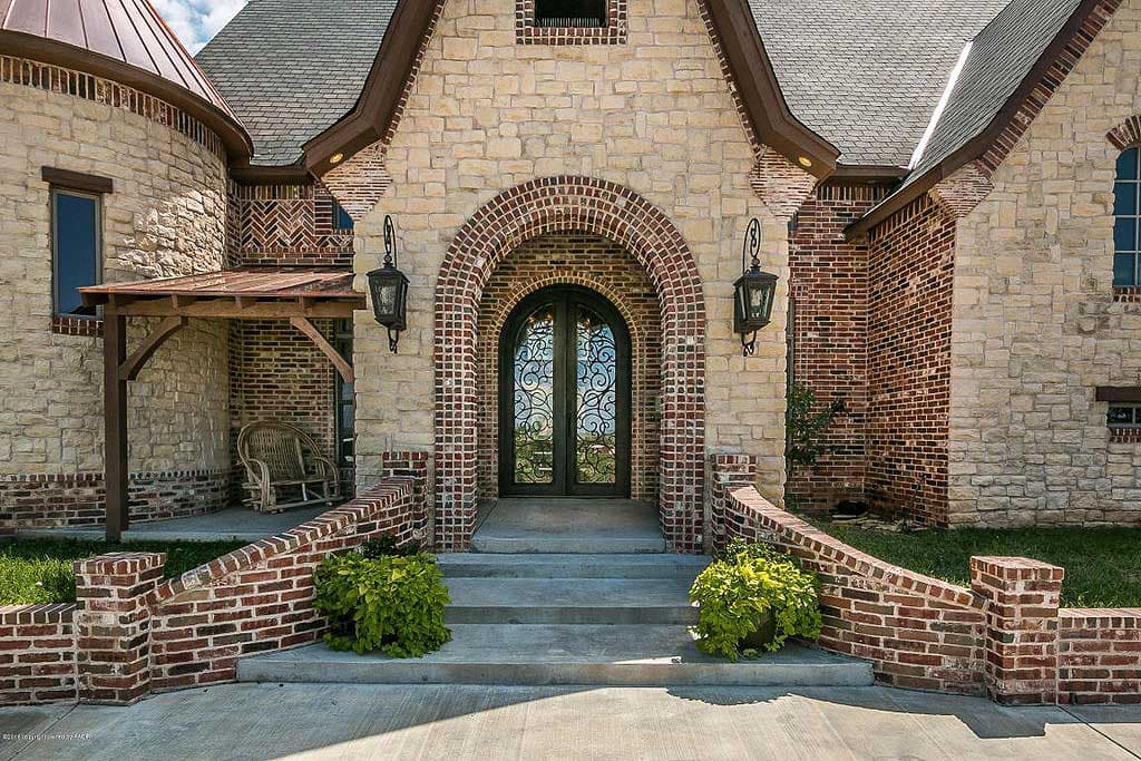 House plan exterior featuring stone and brick facade, arched entry with decorative double doors, covered side porch, and rounded turret.
