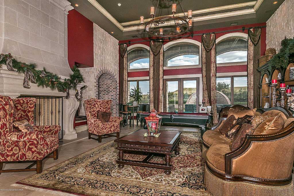 Living room with large stone fireplace, arched windows, patterned armchairs, billiard table, and ornate chandelier.