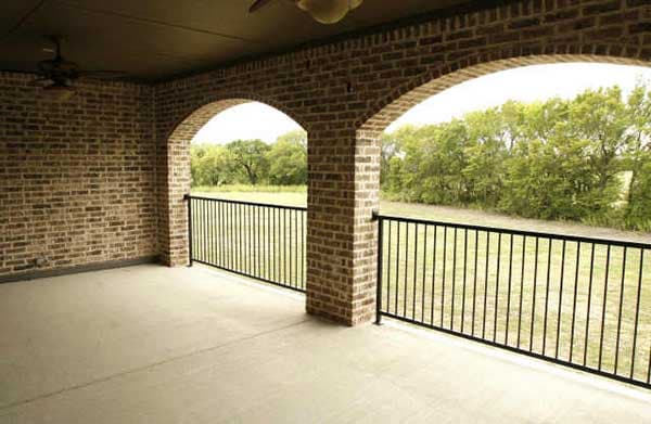 Covered porch with brick columns and two arched openings, featuring a metal railing overlooking a grassy field.