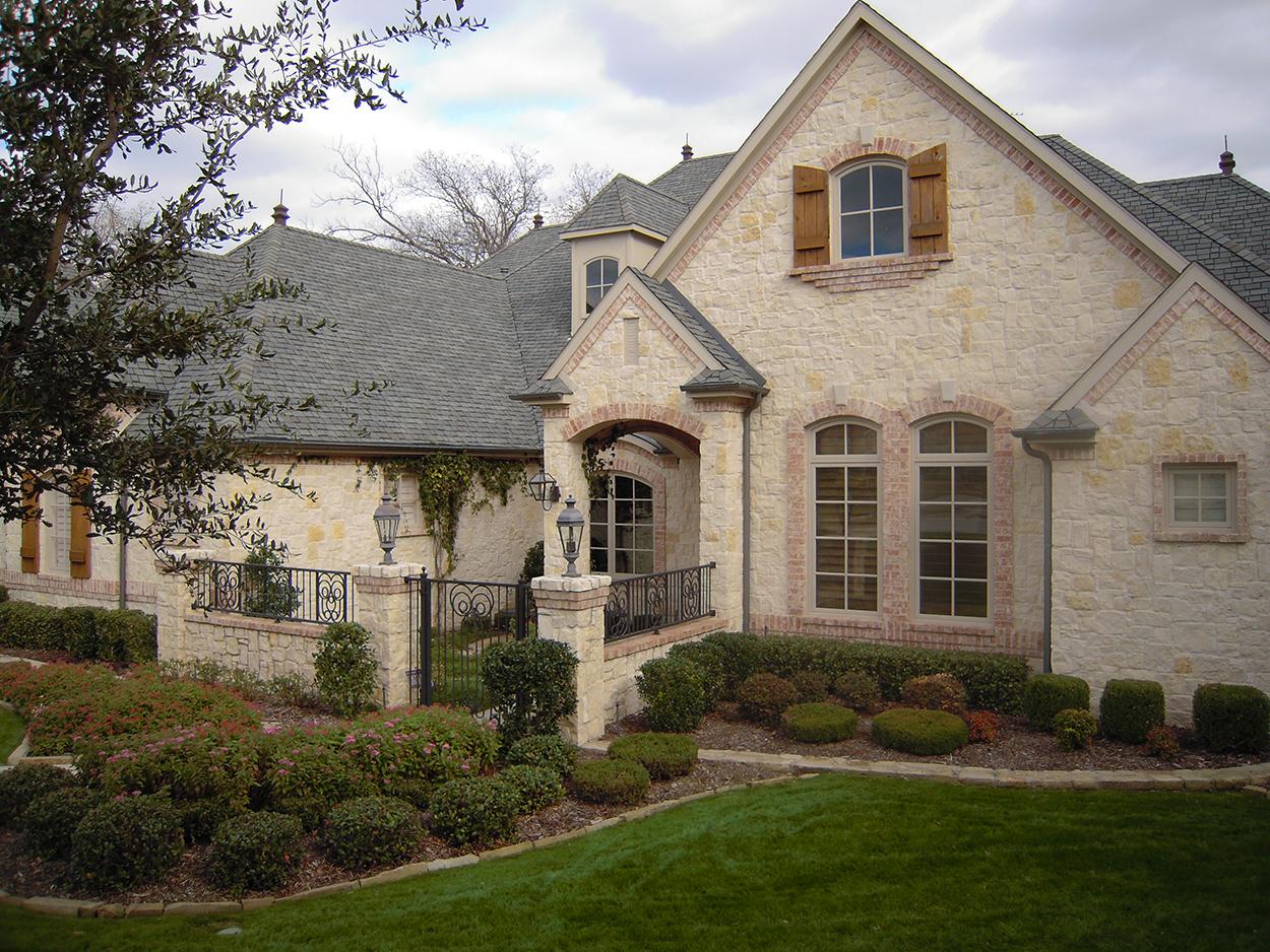 House plan exterior showing stone facade, arched windows with shutters, and a covered entryway with a decorative metal gate.
