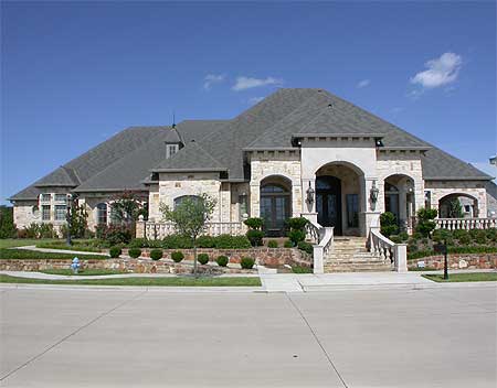 House plan exterior of a large, one-story home with stone facade, arched entryway, multi-gabled roof, and dual garage.