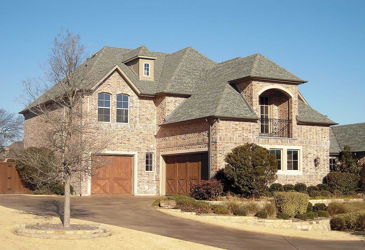 House plan exterior showing a two-story brick home with a dormer, arched balcony, and attached garage.