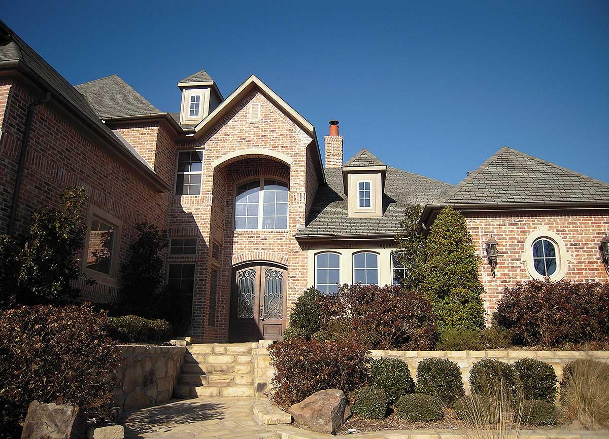 House plan exterior: Two-story brick home with arched entryway, prominent gables, dormers, and stone landscaping.