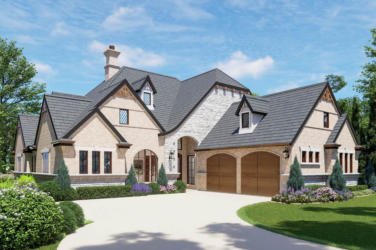 A large, multi-gabled house with tan brick exterior and a curved driveway. Two arched garage doors and small dormer windows add to the upscale design.