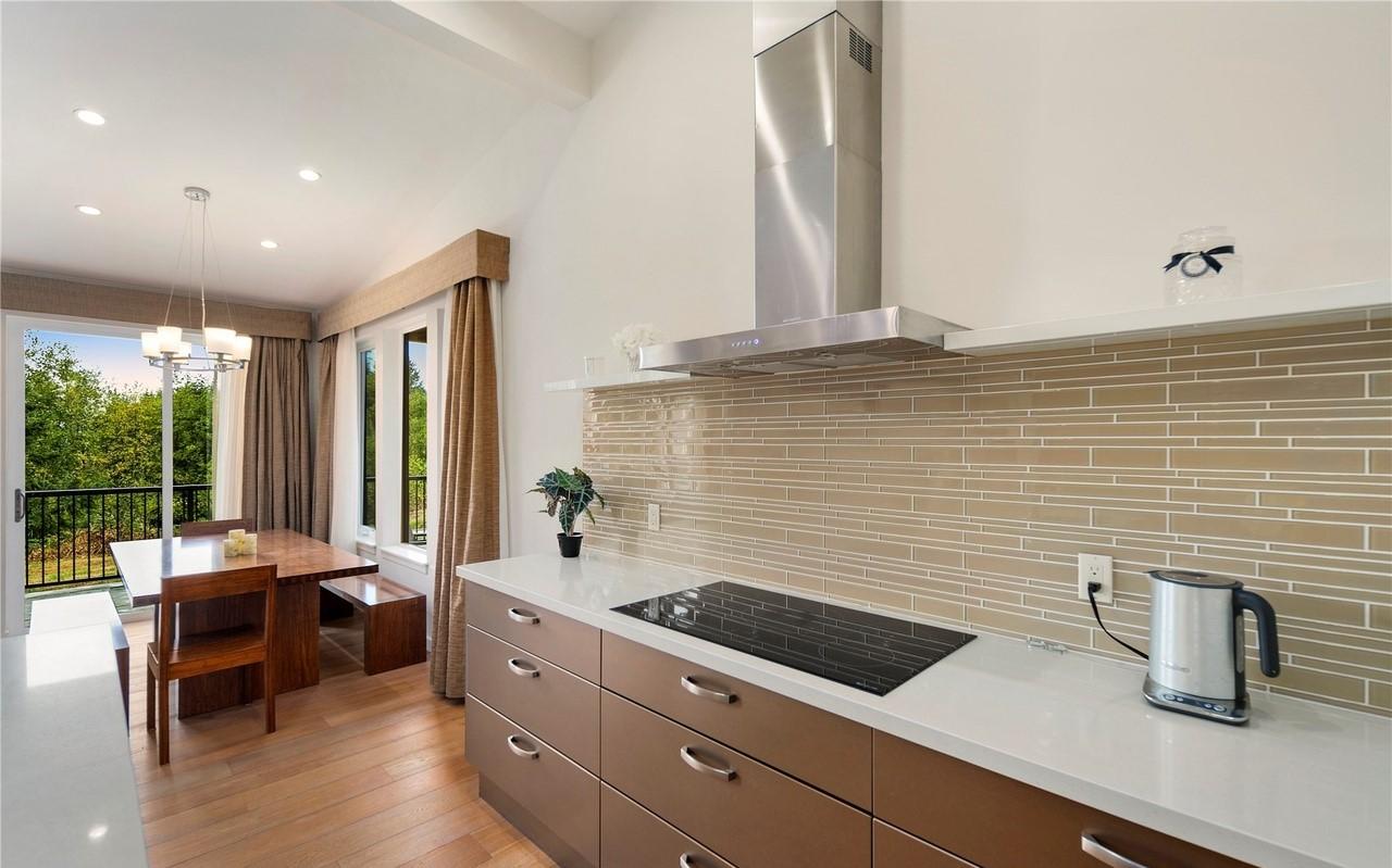 Modern kitchen and dining area with vaulted ceiling, stainless steel range hood, and large windows overlooking trees.
