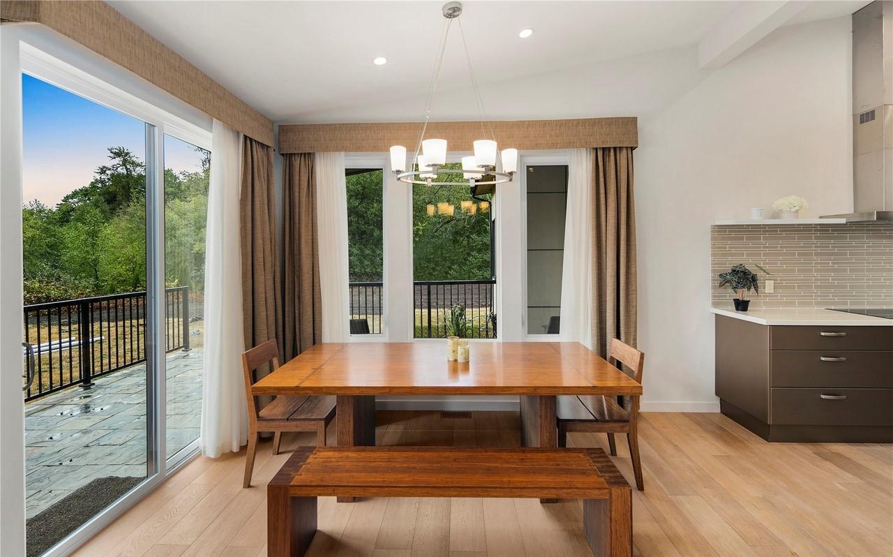 Dining area with large wooden table and benches, modern chandelier, and adjacent kitchen counter with tiled backsplash.